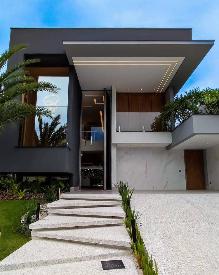 Fachada de casa moderna, com uma estrutura angular e escura, possui janelas grandes e está cercada por vegetação. Há uma entrada elevada com vários degraus que leva à porta da frente. O piso inferior é predominantemente um espaço aberto que serve como garagem para um carro.