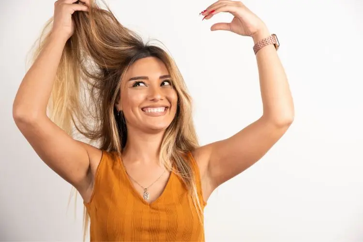 Mulher de pele clara e cabelo loiro escuro passando a mão nos fios e sorrindo
