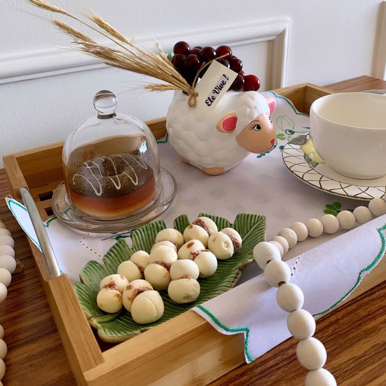 Bandeja de madeira com decoração temática: mini ovelha com tag "Ele Vive!" segurando trigo e uvas, cúpula de vidro cobrindo sobremesa, prato de folhas com biscoitos brancos, xícara em pires decorado e contas brancas complementam o arranjo.