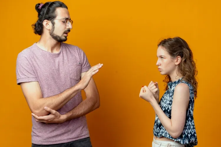 Foto de fundo amarelo com homem de pele clara e cabelo longo preso fazendo gesto para mulher se acalmar e mulher de pele clara com semblante bravo e mãos com punho fechado