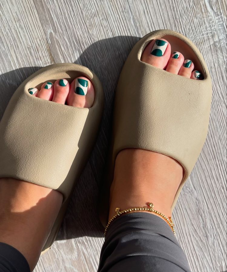 Pés em sandálias bege com unhas decoradas em esmalte branco e verde em design geométrico. Tornozeleira dourada complementa o look, enquanto o piso de madeira clara reflete a luz do sol, criando um efeito iluminado e moderno.