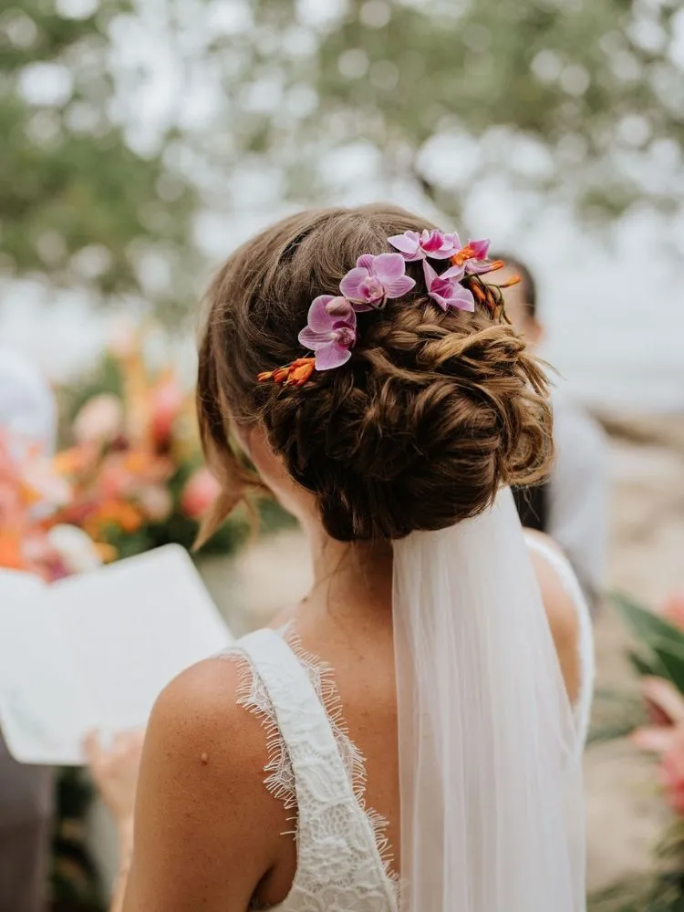 Mulher de cabelo castanho de costas falando votos e usando coque com orquídea lilás