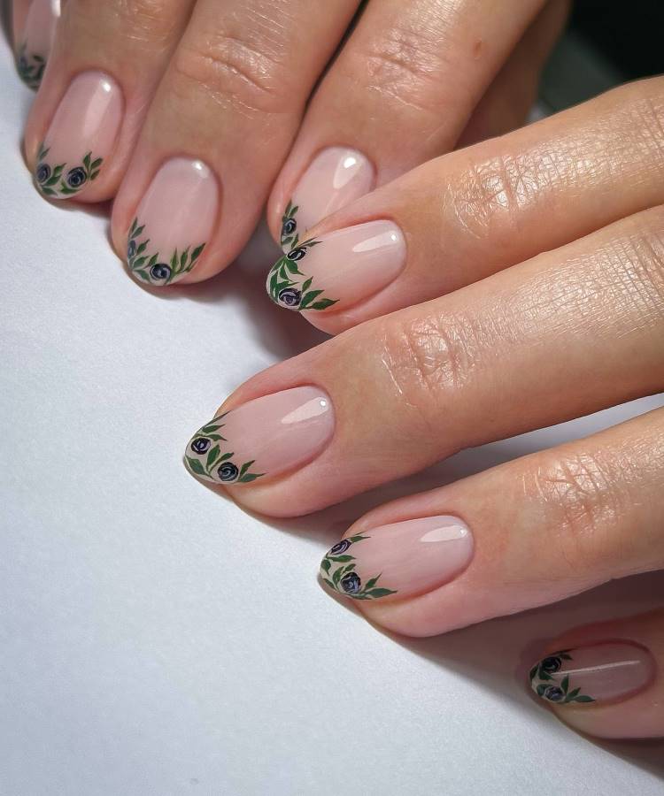 Unhas de gel esmaltadas em tom nude com ponta decorada por delicadas flores negras estilizadas e folhas verdes. O design elegante combina sofisticação e delicadeza, destacando o acabamento brilhante e bem trabalhado sobre o fundo claro.
