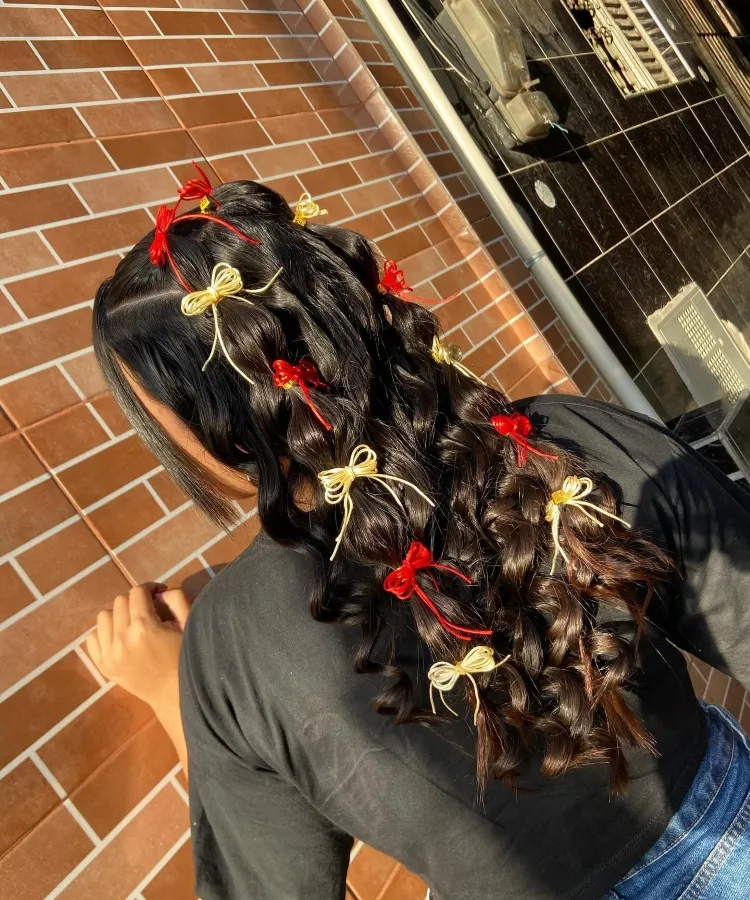 Uma pessoa de pele morena clara, vista de costas, exibe um penteado elaborado com cabelos longos, escuros e ondulados. Pequenos laços vermelhos e dourados estão delicadamente espalhados pelo cabelo. A pessoa veste uma camiseta preta e jeans azuis. Ao fundo, uma parede de tijolos avermelhados e uma janela com luz natural criam um contraste interessante.

