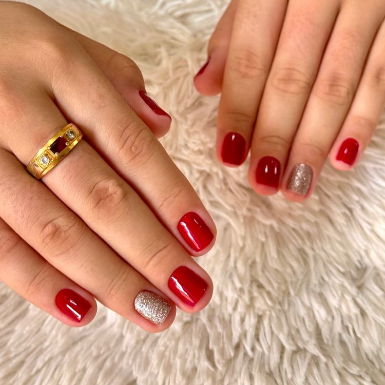 Mãos com estilos de unhas curtas, pintadas em esmalte vermelho vibrante, com destaque para uma unha decorada em glitter prateado. Um anel dourado com detalhes em pedras adorna o dedo. Fundo de tecido branco felpudo, destacando elegância e simplicidade.