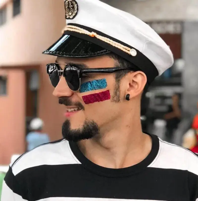 Homem de pele clara com boné da marinha e maquiagem de duas faixas de glitter, uma azul e uma rosa
