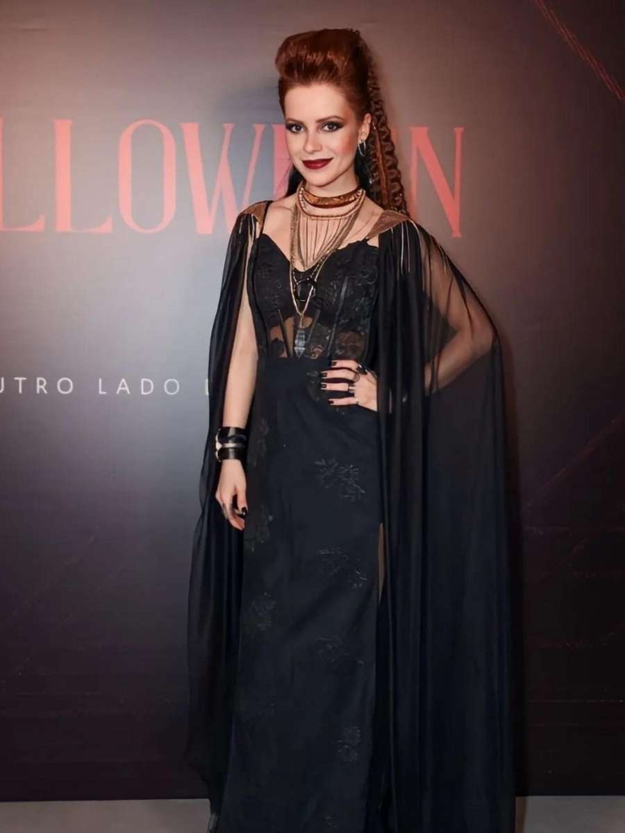 Imagem em fundo preto com palavras escritas. No centro, Sandy com fantasia na festa Halloween da Sephora. Cantora se fantasiou de Bruxa Viking, usando vestido preto com capa preta. Seu penteado é alto e possui tranças.