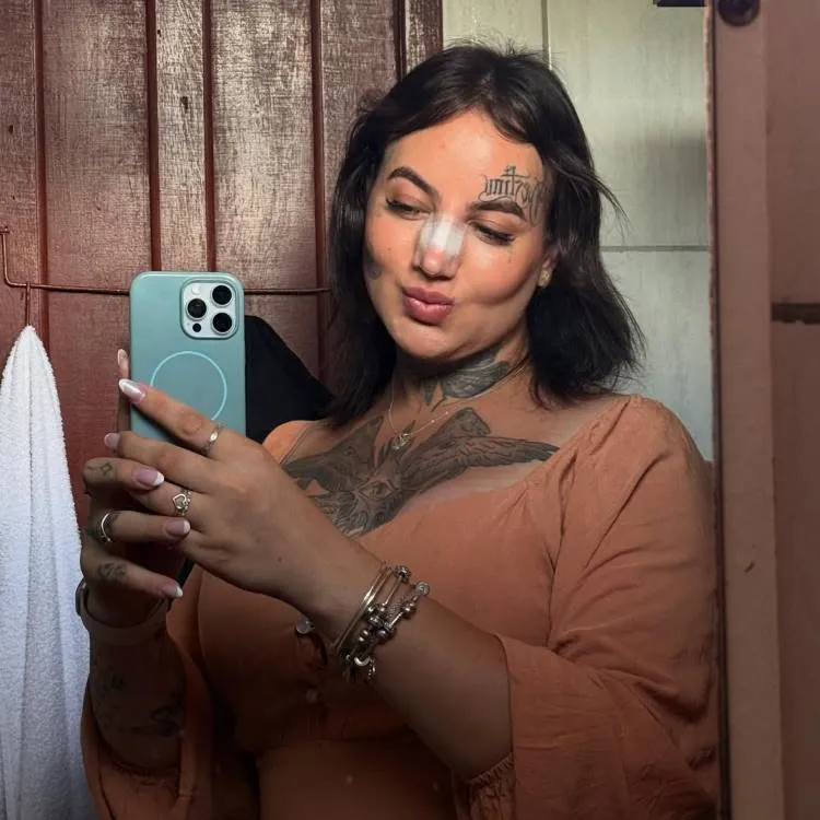 Selfie no espelho de banheiro com paredes de madeira. Mulher de cabelos escuros curtos, tatuagens no peito e rosto, posa com bico nos lábios. Usa blusa marrom de manga longa e exibe curativo branco no nariz após procedimento.