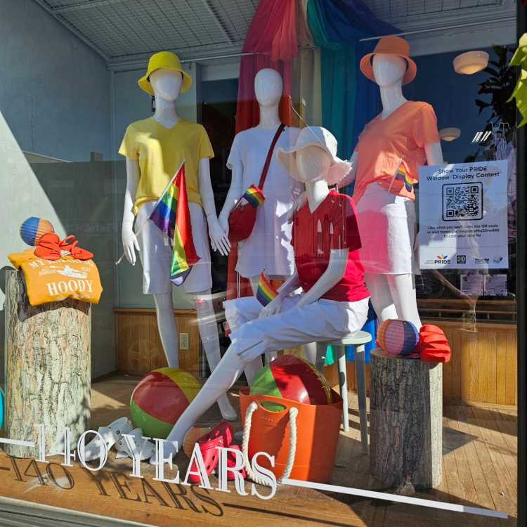 Vitrine decorada com manequins vestindo roupas coloridas em tons vibrantes, com acessórios como chapéus e bolsas. Bandeiras LGBTQIA+ e bolas de praia decoram o ambiente. Fundo destaca tecido arco-íris, celebrando diversidade e orgulho. Letreiro comemora "140 Years".