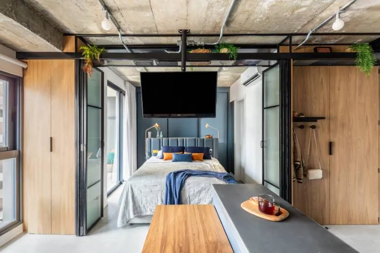 A imagem mostra um quarto moderno com uma estética de design industrial. O quarto possui um teto de concreto exposto com canos e dutos visíveis, pintados de preto. Uma grande cama está posicionada centralmente no espaço, com roupas de cama cinza e almofadas azuis e laranjas que adicionam um toque de cor. Acima da cama, há uma grande TV de tela plana contra uma parede escura, criando um ponto focal. À esquerda da cama, há portas de madeira de altura total, provavelmente armários, que complementam o piso de madeira que se estende por todo o espaço.

À direita do quarto, há uma longa mesa ou bancada de madeira que corre paralela à cama, sobre a qual repousam itens decorativos, incluindo o que parece ser uma bandeja com copo. O quarto é bem iluminado pela luz natural que entra por janelas não visíveis na imagem, mas sugeridas pelos reflexos nas superfícies e objetos dentro do quarto.