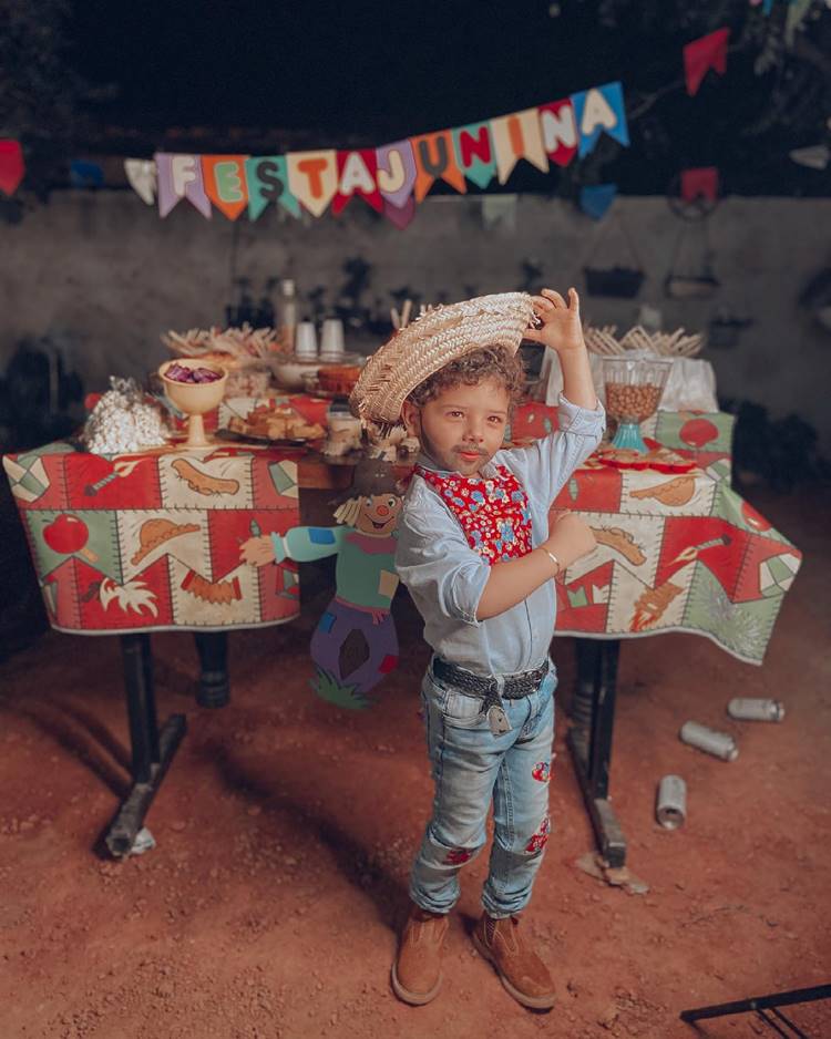 Menino usando traje junino de camisa branca, lenço vermelho, calça jeans, chapéu de palha, em festa junina com mesa repleta de doces