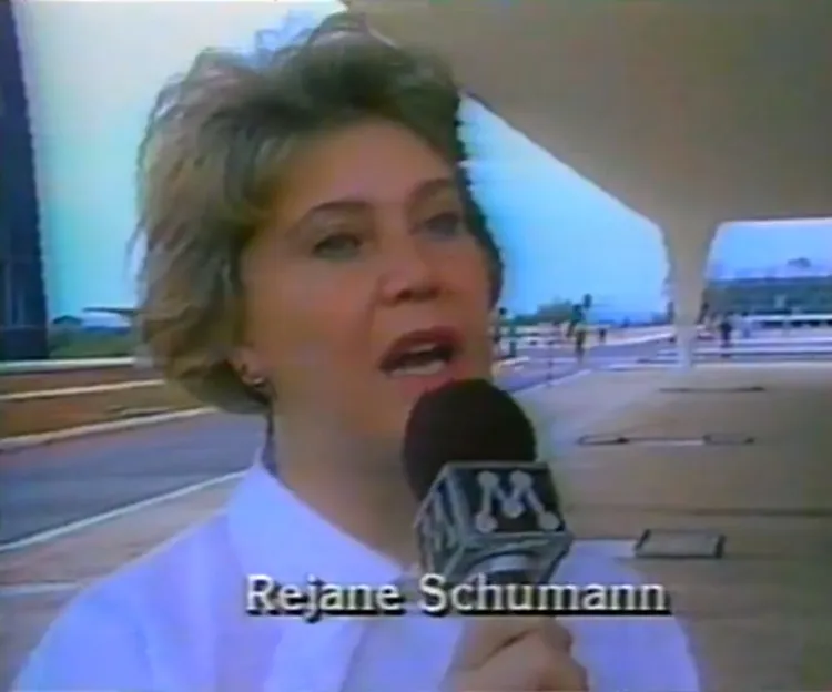 Cena de arquivo televisivo mostrando Rejane Schumann, mulher de cabelos curtos e claros concedendo entrevista em local público. Ela segura microfone com logotipo e legenda na tela exibe o nome “Rejane Schumann”. O fundo revela arquitetura moderna e pessoas circulando.