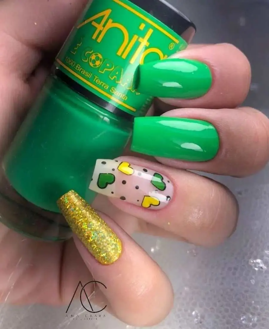 Imagem de mão com unhas decoradas para a Copa do Mundo 2022. Cores usadas, verde, amarelo e glitter dourado. Indicador: verde. Dedo Médio, verde. Anelar: transparente com adesivos de corações verde e amarelo. Mínimo: glitter dourado.