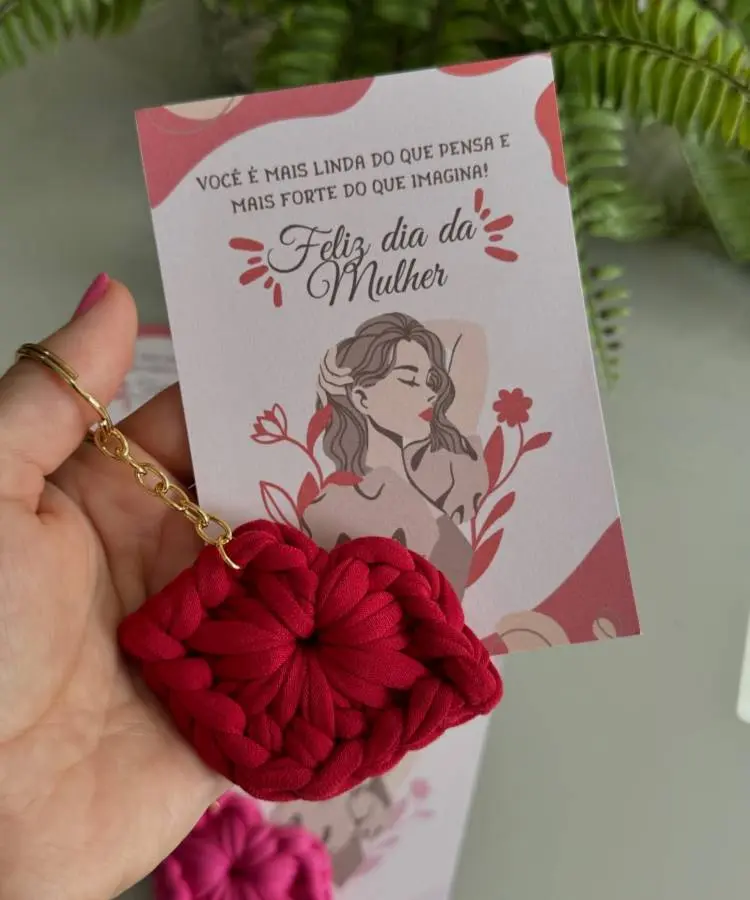 Cartão com a mensagem “Feliz Dia da Mulher” e ilustração feminina em tons vermelhos, acompanhado de chaveiro artesanal em formato de lábios trançados. Fundo decorado com folhas verdes, destacando como presente delicado e criativo para comemorações femininas.
