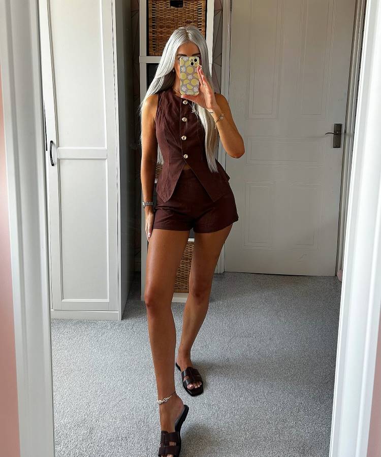 Mulher com cabelos prateados, tirando uma selfie no espelho. Ela usa um conjunto de colete e shorts marrons com botões dourados e sandálias. O fundo mostra um quarto com móveis brancos e um tapete cinza.