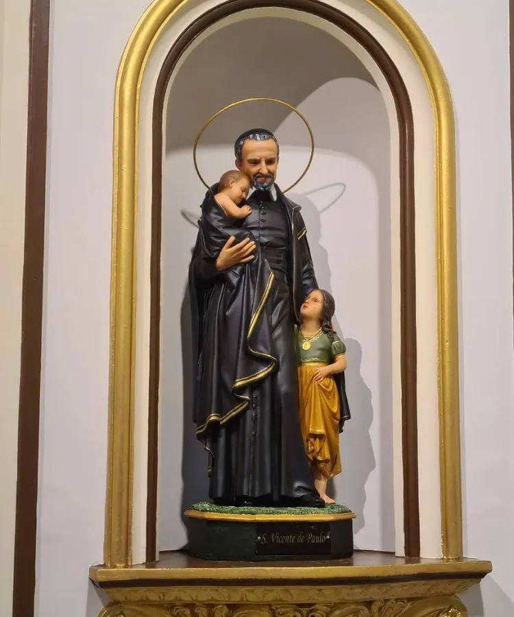 A imagem mostra uma estátua de São Vicente de Paulo em um nicho dourado. Ele veste uma túnica preta e segura um bebê adormecido. Ao seu lado, uma criança olha para ele. Um halo dourado circunda sua cabeça, simbolizando santidade. A base da estátua tem seu nome.


