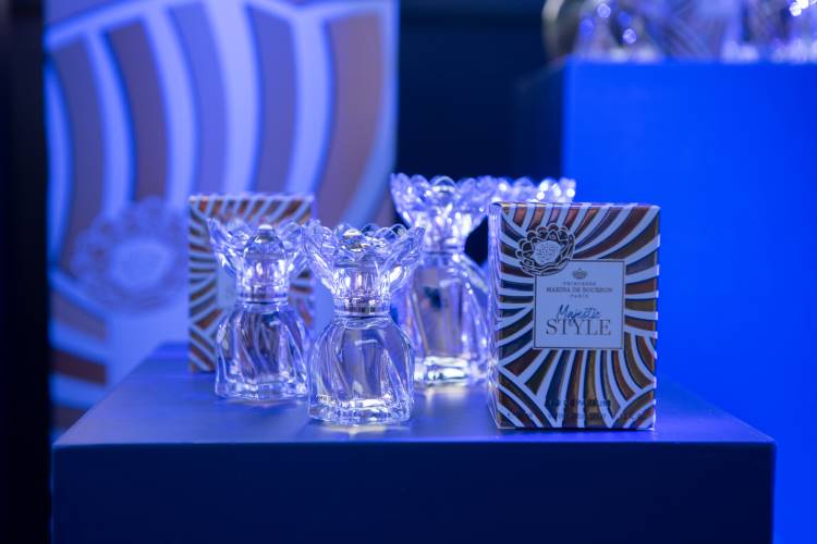 Frascos de perfume cristalinos com tampas decorativas estão dispostos sobre uma mesa azul. Embalagens com design dourado e azul estão ao lado, destacando o nome "Marina de Bourbon". A iluminação azul cria uma atmosfera sofisticada e elegante ao redor dos produtos.