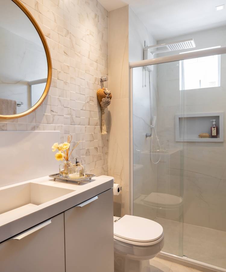 Banheiro decorado em estilo minimalista, com um mosaico de parede em tons claros, espelho redondo dourado, pia com bancada branca e detalhes decorativos. Box de vidro com chuveiro moderno, iluminação natural e design organizado promovem um ambiente limpo e elegante.





