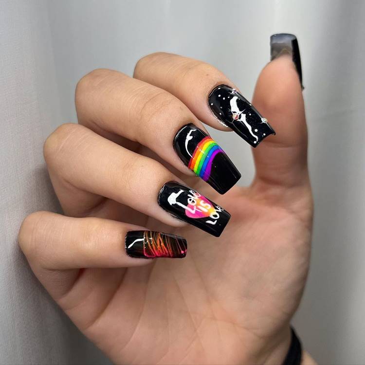 Mão com unhas decoradas coloridas: Polegar: Preto Indicador: Preto com pedrinhas Médio: Preto com arco-íris no meio Anelar: preto com desenho de coração colorido e frase Love is Love Mínimo preto com riscos laranja e rosa