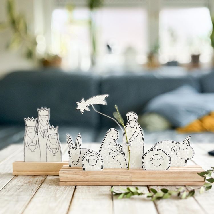 A imagem mostra um presépio minimalista com figuras de papel em bases de madeira. Os personagens incluem Maria, José, o Menino Jesus, os Três Reis Magos e animais. Uma estrela cadente destaca-se acima, enquanto ramos verdes decoram a cena, que está sobre uma superfície de madeira clara.


