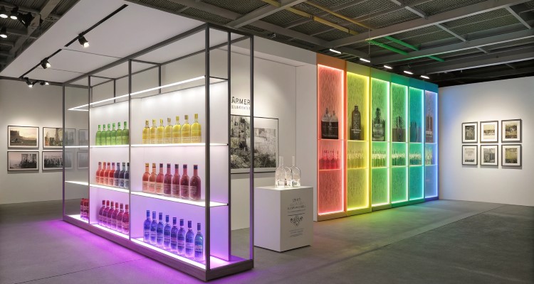 Espaço iluminado com estantes minimalistas exibindo garrafas coloridas organizadas em tons de arco-íris. Parede ao fundo apresenta prateleiras com iluminação LED em cores vibrantes. Fotos em preto e branco decoram a lateral. Ambiente clean, moderno e inclusivo.
