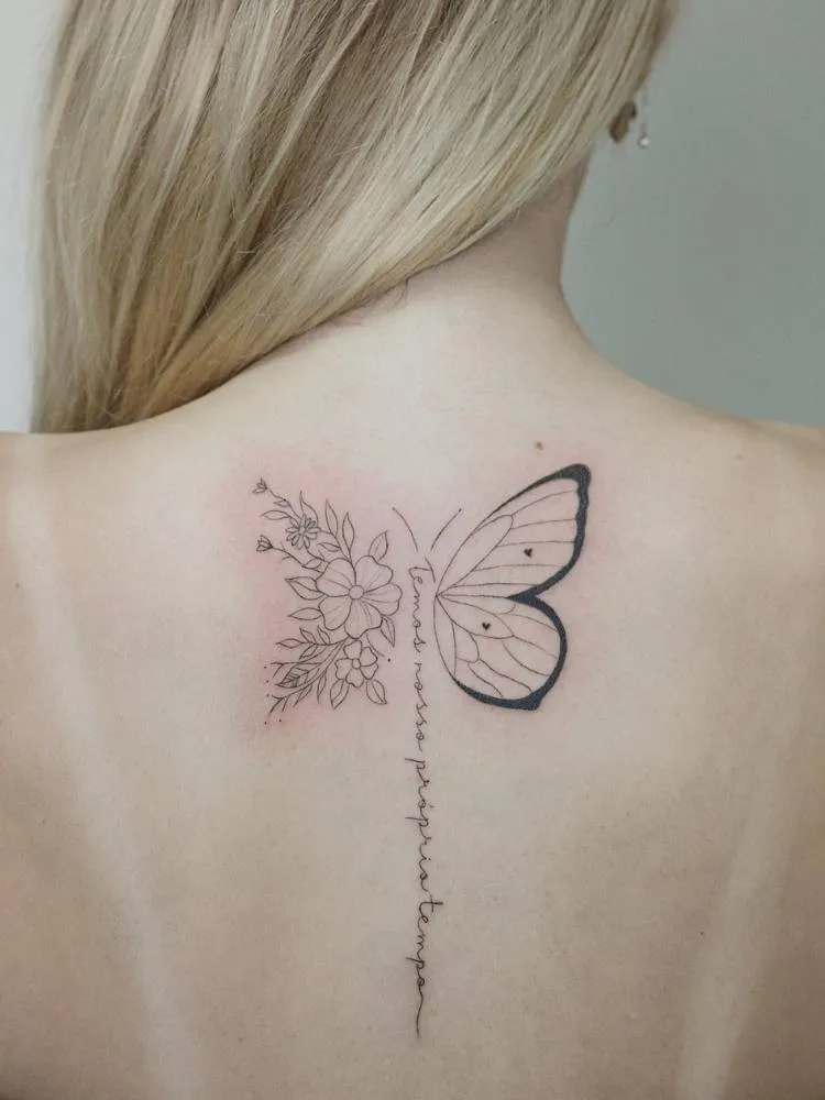 Tatuagem de borboleta com metade da asa em traços finos e grossos. A outra metade da asa é comporta por flores. O corpo do inseto é uma frase que se estende na coluna: "temos nosso prórpio tempo". Ela foi feita nas costas de uma mulher loira de pele clara.