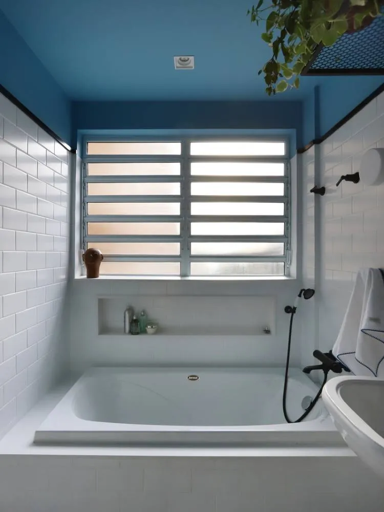 Esta imagem exibe um banheiro com um teto colorido azul-claro. Uma banheira branca retangular com nicho embutido ocupa o centro, sob uma janela com persianas horizontais. As paredes são revestidas com azulejos brancos tipo metrô, e plantas penduradas complementam a decoração.

