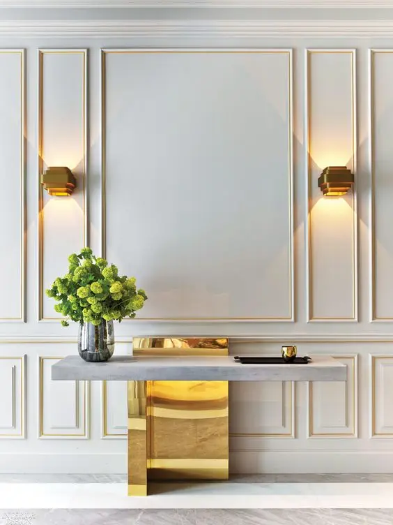 Hall de entrada com aparador cinza e dourado, vaso com plantas e paredes brancas com boiserie branco e dourado