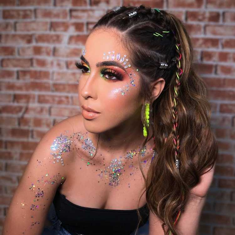 Mulher de pele clara com cabelo semi-preso, maquiagem neon com glitter e pedrarias