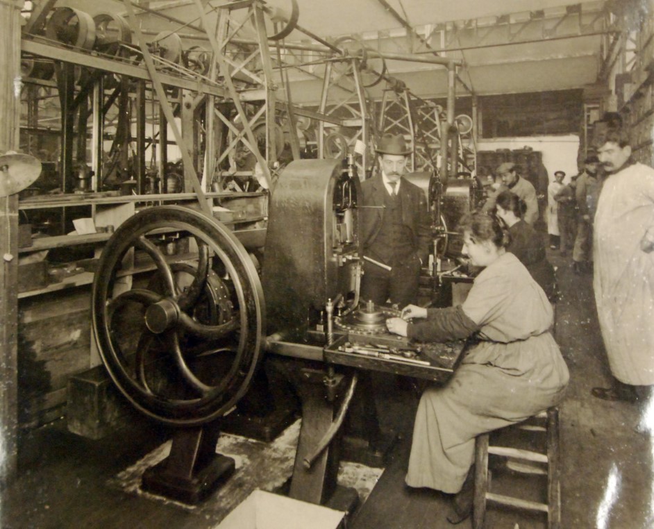 Foto em preto e branco do interior de uma fábrica de botões com uma mulheres sentadas a operar máquina sob a supervisão de homens ao redor