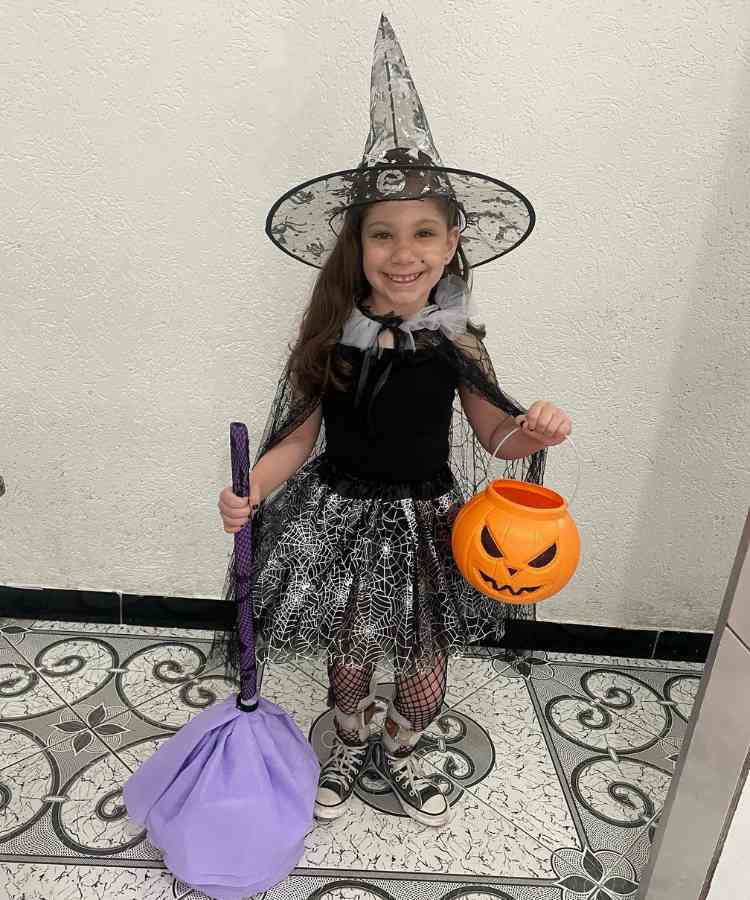 Garota com fantasia de bruxa infantil. Cores predominantes: preto e prata. Usa: chapéu pontudo, capa, body, saia, meia calça arrastão, tênis, segura vassoura de brinquedo e baldinho formato cabeça de abóbora