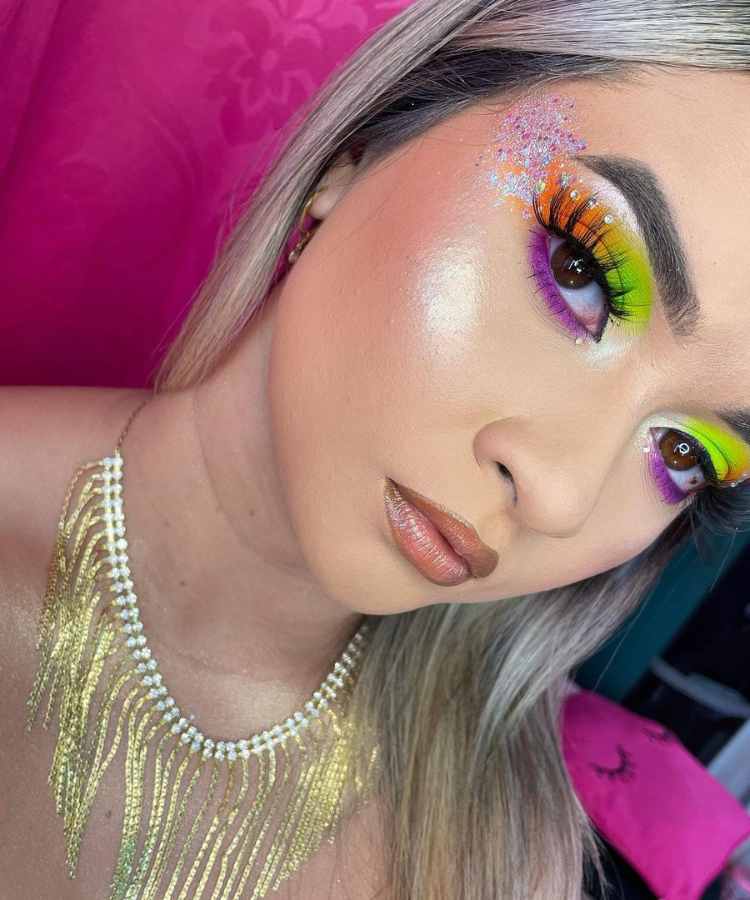 Mulher de pele clara com pele glow, cílios postiços, maquiagem neon em laranja, verde  e roxo e glitter