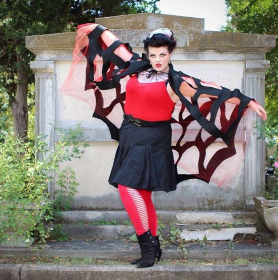 Foto de mulher com capa de aranha e tule vermelho por baixo, blusa vermelha, saia preta no joelho e meia-calça vermelha vazada