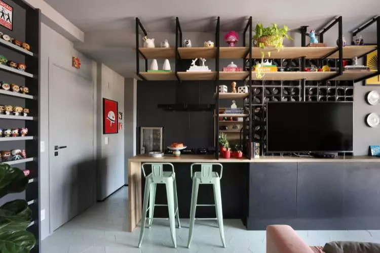 Estante vazada dividindo sala e cozinha mas sem separá-los totalmente, com TV, vasos e diversos itens decorativos. Ainda, ao lado, há prateleiras que armazenam bonecos temáticos. 
