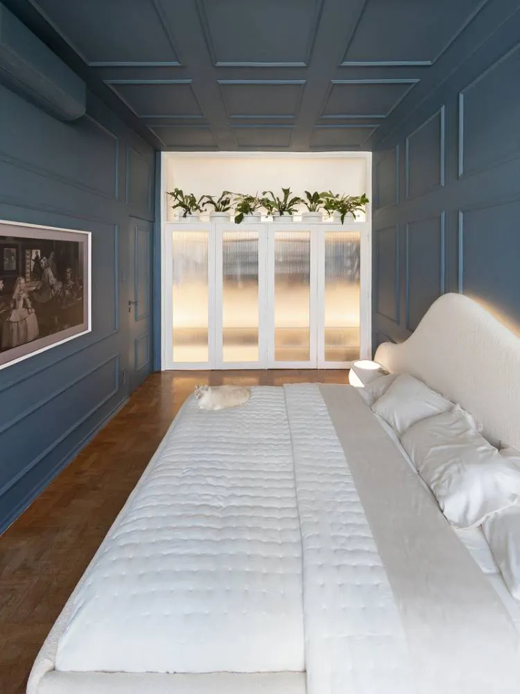 Esta é uma imagem de um quarto elegantemente decorado. Uma cama grande com roupa de cama branca domina o centro, com um gato creme deitado nela. As paredes e o teto são de um azul-petróleo profundo com painéis. Ao fundo, portas de vidro fosco com plantas em cima adicionam um toque de natureza.

