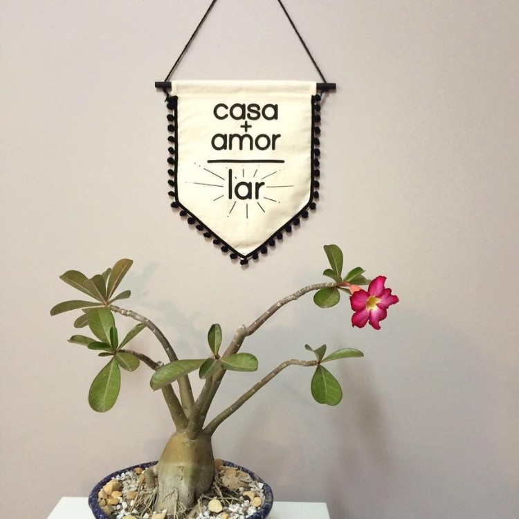 Vaso com rosa do deserto sob aparador branco, com flâmula bege escrito casa + amor = lar