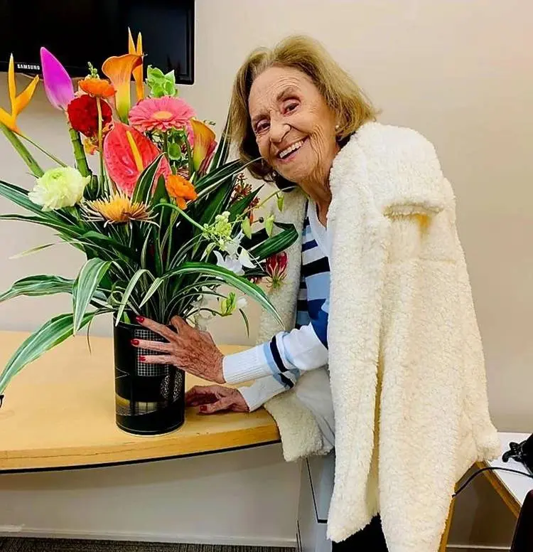 Laura Cardoso hoje em dia, em foto sorrindo e segurando vaso com flores coloridas.
