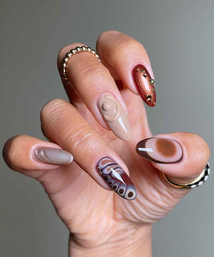 Unhas decoradas com design variado: uma em nude com relevo, outra marrom com pedras douradas, uma com padrão abstrato em tons marrons, e duas em nude e marrom com detalhes ondulados. O fundo é cinza, destacando a arte criativa e elegante.