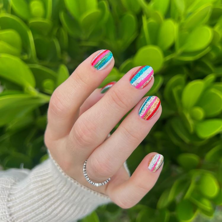 Mão destacando estilos de unhas curtas, decoradas com listras verticais coloridas e brilhantes em tons de azul, rosa, dourado e verde. A pele clara contrasta com o fundo vibrante de folhas verdes, criando um visual alegre e moderno.