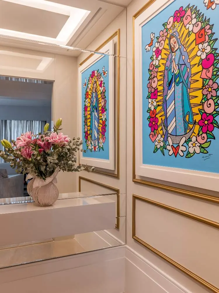 Hall de entrada com espelho, aparador branco, vaso com flores rosas, parede off white com boiserie dourado e quadro colorido 
