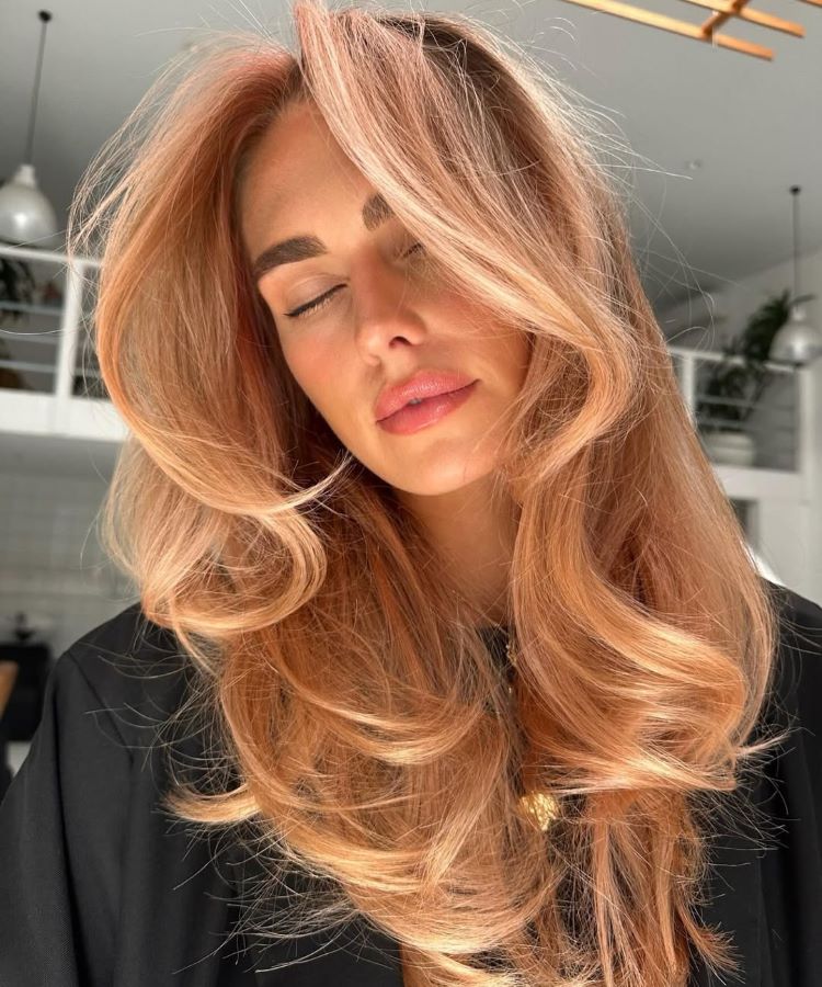 Mulher com cabelos longos e volumosos, destacando cores de cabelo tendência como rosé gold, com tons suaves e brilhantes. Ela veste roupa preta, usa maquiagem leve com lábios rosa claros e está em ambiente interno iluminado por luz natural.
