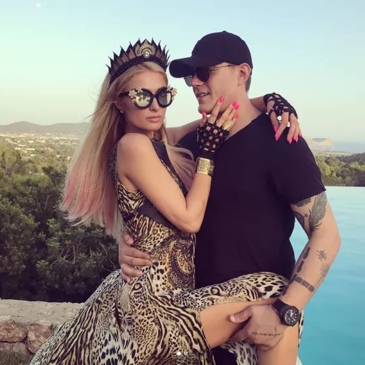 Paris Hilton e Chris Zylka posando ao ar livre; a mulher, usando vestido de estampa animal, coroa, óculos decorativos e luvas, está nos braços do homem, que veste boné, camiseta preta e óculos escuros; fundo natural com árvores e piscina.