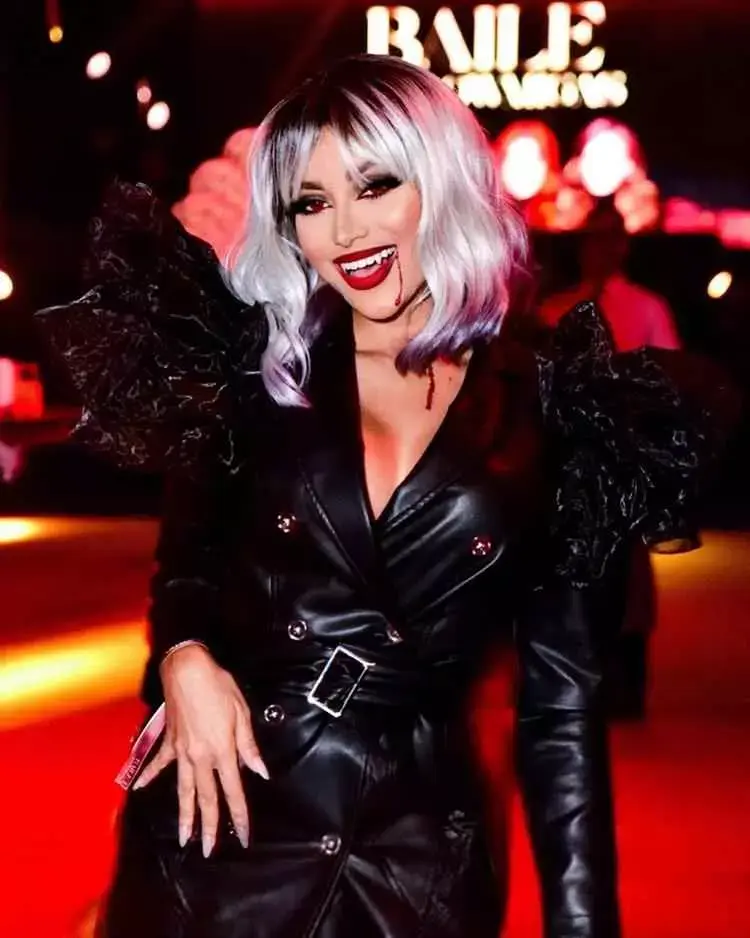 Mulher de pele clara com cabelo platinado, lente vermelha, batom vermelho, dentadura de vampira e sangue falso