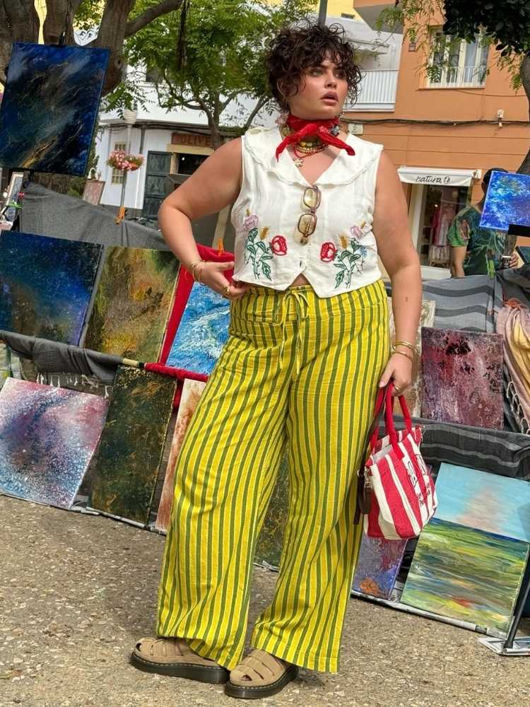 Mulher de pele clara usando colete branco bordado, calça verde e preta listrada e bandana vermelha no pescoço