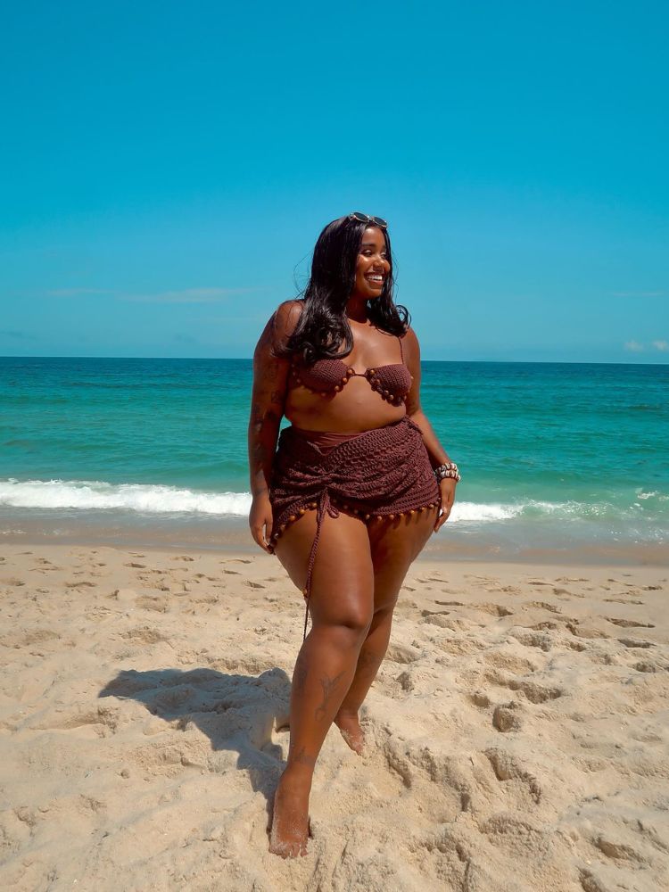 Mulher de pele negra na praia usando biquini e saida de praia marrom em crochê