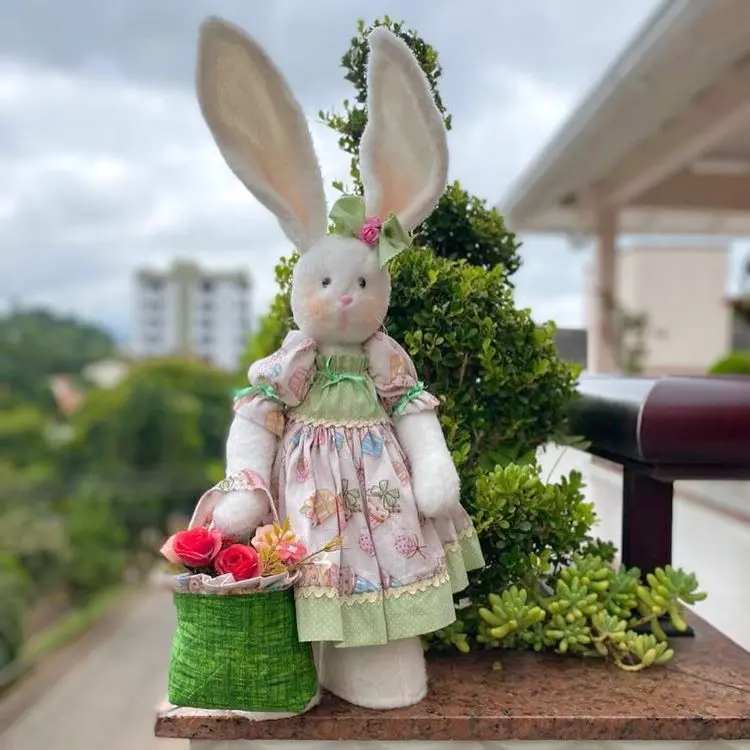 Coelha de tecido branco com vestido floral em tons verde e rosa, laço na cabeça e cesta verde com flores artificiais. Cenário ao ar livre sobre superfície de pedra, com arbustos, plantas decorativas e edifícios ao fundo.