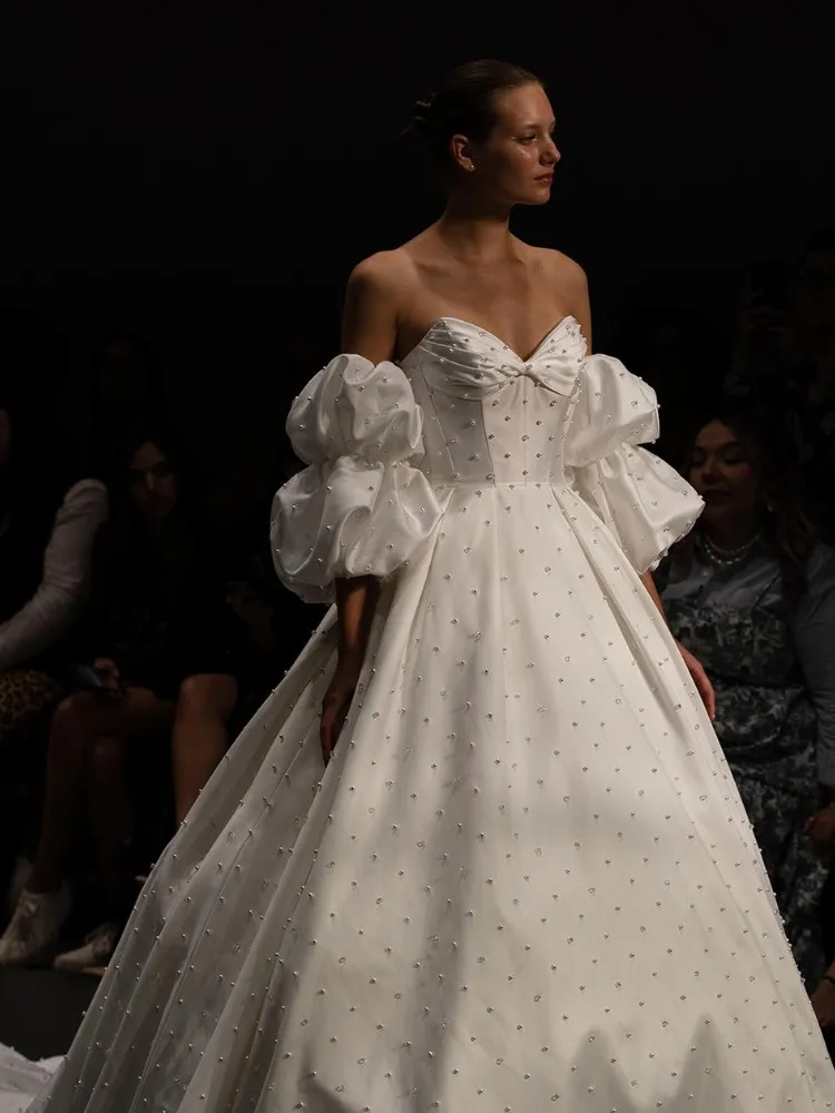 Mulher de pele clara desfilando com vestido de noiva sem alça, mangas bufantes e todo bordado em pérola