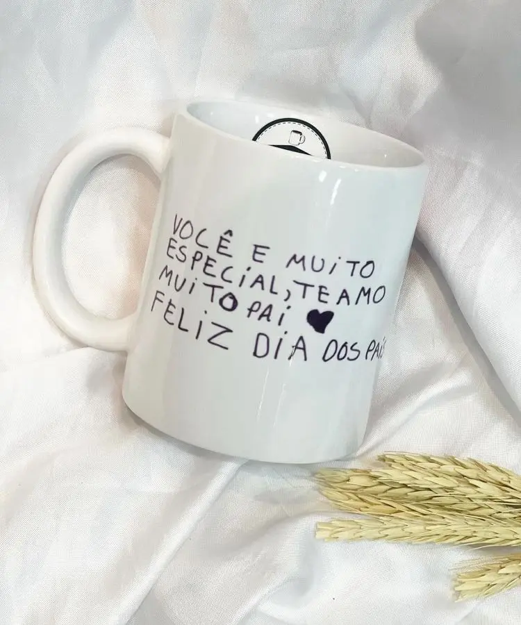 Uma caneca branca com texto manuscrito e um desenho de coração, sobre um tecido branco e ao lado de espigas secas.