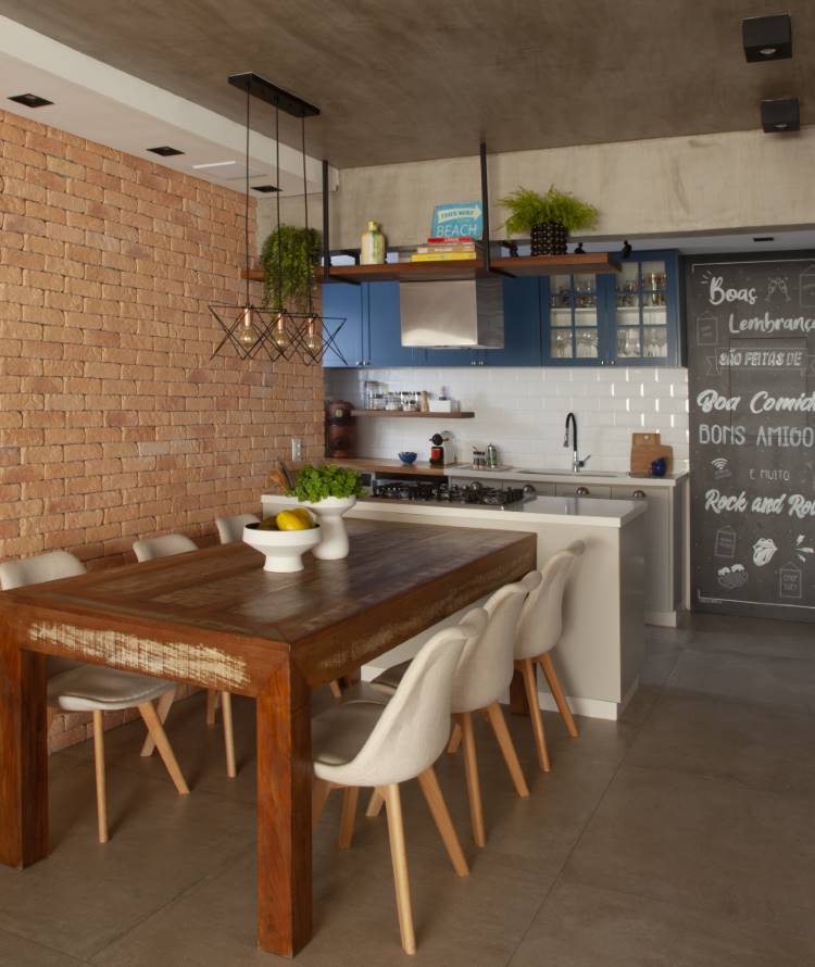 Cozinha e sala de jantar integradas com parede de tijolinhos e teto de cimento queimado. A mesa de madeira rústica com cadeiras brancas modernas contrasta com a cozinha de armários azuis, criando um ambiente contemporâneo.







