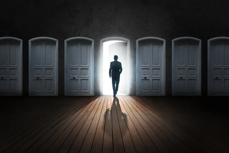 Homem caminha em direção a uma porta iluminada, enquanto outras permanecem fechadas em um corredor escuro. A imagem simboliza escolhas estratégicas e a capacidade de "transformar crise em oportunidade" ao encontrar novas possibilidades e soluções em momentos difíceis.
